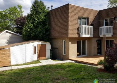 L’extérieur de la maison a été grandement amélioré. Dans un premier temps, la porte d’entrée a été remplacée par une porte noire contemporaine à panneaux givrés. Les balustrades des balcons ont été repeintes. Une très grande terrasse de bois a été ajoutée. Cette dernière permettra facilement d’y installer une table de belle grandeur ou autre ameublement d’extérieur. Une allée de tuiles de pierre permet maintenant une circulation beaucoup plus confortable entre l’entrée de la cour et le cabanon et la terrasse. Pour terminer, une très belle palissade de bois rend désormais la cour beaucoup plus intime.