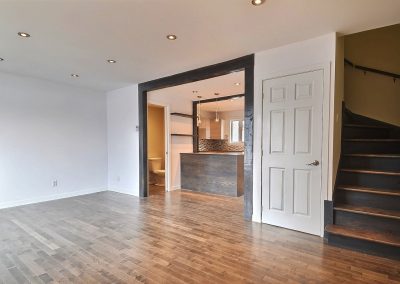 La transition entre le salon et la cuisine à aire ouverte est mise en valeur par les deux arches parallèles ornementées de poutres teintes décoratives.   Le plancher du salon en bois franc enrichit la pièce et y ajoute de la chaleur. La teinture de l’escalier permet un agencement avec le plancher du salon.